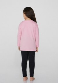 Světle růžová dlouhá rukávová košile s kulatým výstřihem, kombinovaná s černými legínami, které mají hladkou texturu a volný střih.