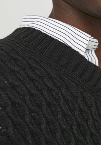 Black knit sweater with a textured cable pattern, featuring a ribbed neckline. A white collared shirt with blue stripes is visible underneath.