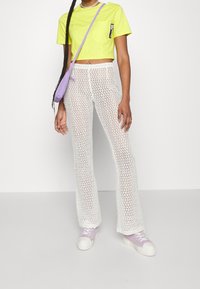 Ljusgul kort t-shirt med ficka, vita virkade utsvängda byxor och lavendelfärgad axelväska. Modell som bär ljuslila sneakers.