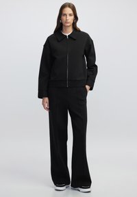 Zwarte zip-up jas met een kraaghalslijn, gecombineerd met bijpassende wide-leg broeken. Glad materiaal met een ontspannen pasvorm en minimale details.