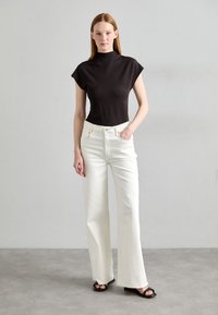 Haut noir à col montant avec des manches courtes associé à un jean large blanc taille haute. Le mannequin porte des sandales noires. Look simple, épuré et moderne.