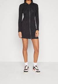 Vestido negro de manga larga con collar y cremallera frontal, con un diseño ajustado y un sutil patrón texturizado, combinado con zapatillas blancas y negras.