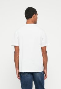 Hombre de espaldas que lleva una camiseta blanca de manga corta y jeans de denim azul, contra un fondo blanco liso.