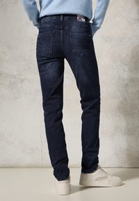 Dark blue denim jeans with a slim fit, featuring two back pockets and a waistband label, paired with light-colored sneakers.