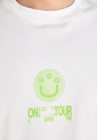 Biała bawełniana koszulka z zieloną grafiką, w tym uśmiechniętą buźką oraz tekstem "ON TOUR SUPPORT FROM", z teksturowanym wzorem.