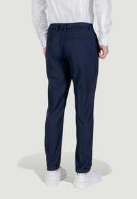Homme portant un pantalon sur mesure bleu marine, une chemise blanche à boutons, et des baskets blanches, vu de dos sur fond blanc.