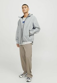 Hellgraue Jacke mit einem strukturierten Muster, schwarzen Akzenten, Kapuze und elastischen Bündchen. Kombiniert mit beigen Hosen und mehrfarbigen Turnschuhen.