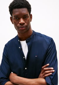 Camisa azul marino abotonada con cuello mao, tela ligera y un pequeño detalle de logo. Llevada sobre una camiseta blanca de cuello redondo.