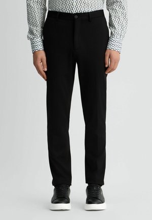 Schwarze Hose aus glattem Stoff, mit Slim-Fit, Gürtelschlaufen, einem Ein-Knopf-Verschluss und kombiniert mit schwarzen Sneakers.