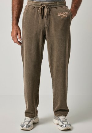 Pantaloni della tuta marroni con coulisse con testo UNLMTD 2014 sull'anca sinistra, indossati da una persona con le mani vicino alle tasche e scarpe da ginnastica bianche-grigie.