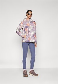 Chaqueta morada con patrón floral y acentos naranjas, combinada con leggings azules y zapatillas marrones. Cuenta con capucha y cierre de cremallera.