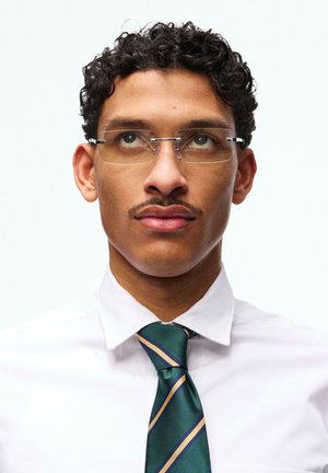 Jonge man met krullend haar, zonder montuur bril, wit overhemd en groene streepvormige das, die omhoog kijkt tegen een effen achtergrond.