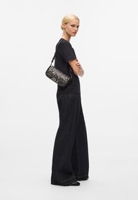 Schwarze Handtasche mit einem strukturierten, glitzernden Design, die über die Schulter getragen wird. Modell in schwarzem Shirt und weit geschnittenen, dunklen Jeanshosen, seitlich blickend.