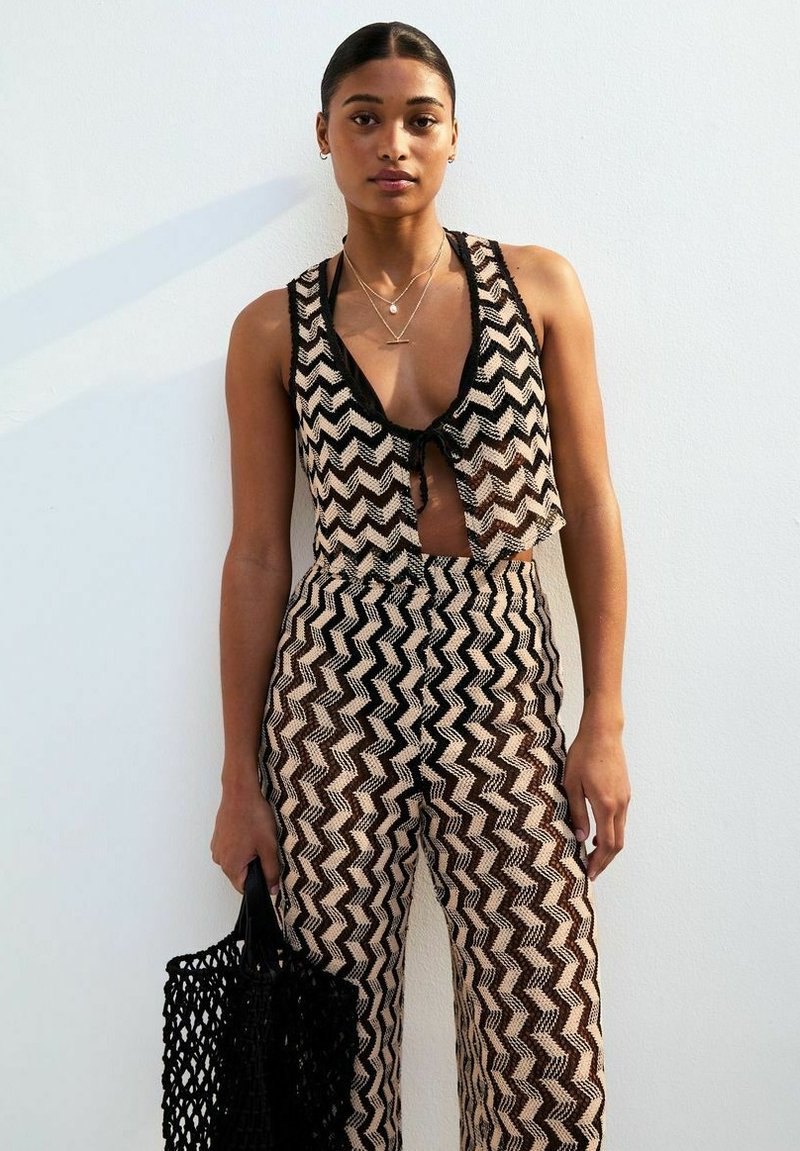 Haut à crop en crochet avec un pantalon assorti noir et beige à motif zigzag. Les accessoires incluent des colliers superposés et un sac fourre-tout tissé noir.