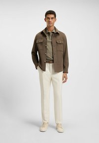 Bruine button-up jas met twee borstzakken, olijfgroene polo eronder, crèmekleurige broek en beige sneakers. Glad materiaal en op maat gemaakte pasvorm.