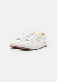 Fehér sneaker, sárga kiegészítőkkel, bőrből és textíliából készült. Kerek orrú, fűzős kialakítás, kaucsuk talp, amelynek felszíne texturált tapadást biztosít.