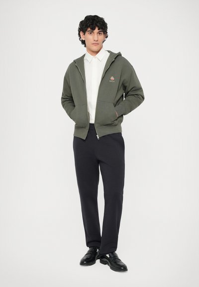 Joven de pie con las manos en los bolsillos, vistiendo una sudadera olive verde con cremallera, una camisa blanca con cuello, pantalones negros y zapatos negros.