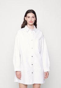 Vestido de camisa em denim branco com mangas longas, fecho de botão na frente e bolso no peito. Apresenta um corte descontraído e detalhe de bainha desfiada.