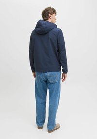 Marineblauer Kapuzenjacke mit glatter Textur, kombiniert mit hellblauen, locker sitzenden Jeans. Das Model steht vor einem hellen Hintergrund.