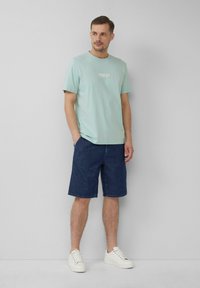 Błękitny bawełniany t-shirt z logo, zestawiony z ciemnoniebieskimi szortami dżinsowymi i białymi trampkami. Model stoi z rękami w kieszeniach szortów na szarym tle.