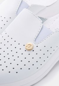 Vita slip-on skor med perforerat mönster, ljusblå detaljer, gyllene logotyp och en strukturerad vit sula.