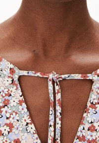 Blouse à motif floral avec un col en V et une fermeture à nouer. Présente de petites fleurs blanches et bleues sur un fond rouge et vert. Texture du tissu douce.