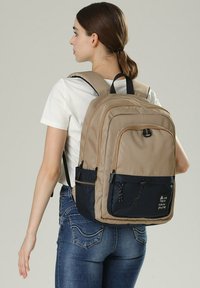 Zaino beige e blu navy con superficie texturizzata, scomparti multipli, spallacci regolabili e una toppa con logo frontale.