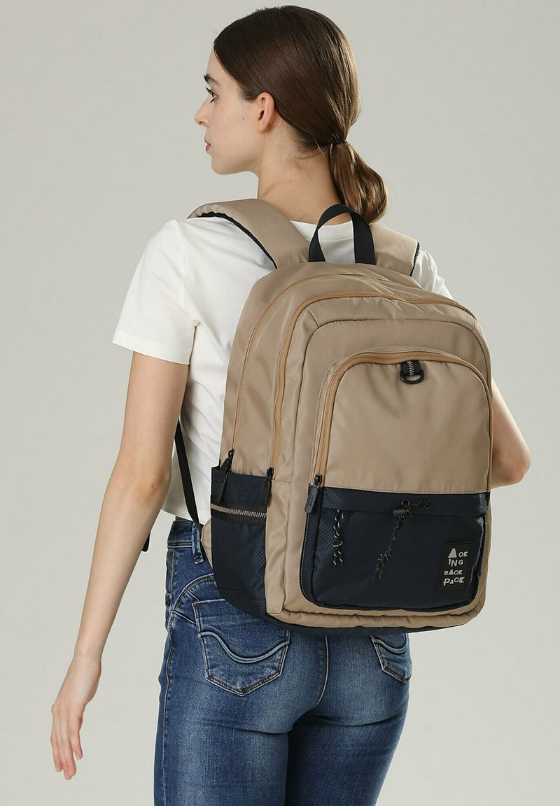 Zaino beige e blu navy con superficie texturizzata, scomparti multipli, spallacci regolabili e una toppa con logo frontale.
