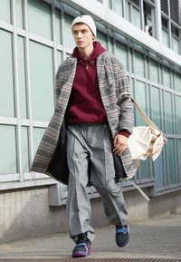 Jeune homme marchant dehors portant un bonnet blanc, un manteau à carreaux, un sweat à capuche bordeaux, un pantalon gris à carreaux et des baskets Nike noir-violet, portant un sac blanc.