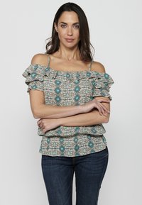 Blusa off-the-shoulder con stampa floreale, maniche a volant, dettagli turchesi e busto aderente, abbinata a jeans scuri.