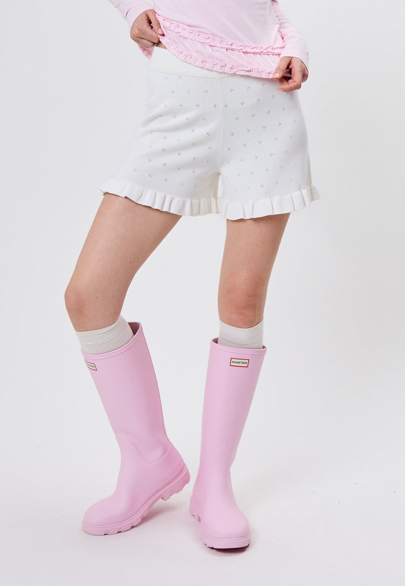 Personne portant un short blanc à œillets avec ourlet à volants, un haut rose clair à volants, et des bottes de pluie Hunter rose pastel montant jusqu'aux genoux.