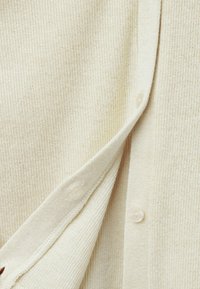 Kremowy kardigan o fakturze prążkowanej, z okrągłymi guzikami z pereł, miękkim splotem i delikatnym połyskiem. Widoczne detale przeszycia.