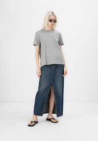 T-shirt gris à manches courtes associé à une jupe maxi en denim bleu à fente haute. Mannequin portant des lunettes de soleil noires et des tongs noires.