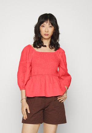 Top peplum de linho vermelho com mangas bufantes, decote quadrado e corpete franzido. Usado com calções castanhos, o modelo está de pé contra um fundo simples.