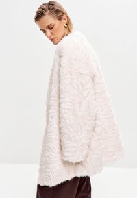 Abrigo de pelo sintético en rosa suave con un aspecto texturizado y esponjoso. Diseño oversized y mangas anchas, combinado con pantalones marrones.