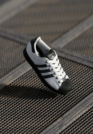 Biało-czarne buty sportowe Adidas z wzmocnionym noskiem i trzema paskami, umieszczone na teksturowanej metalowej kratce.