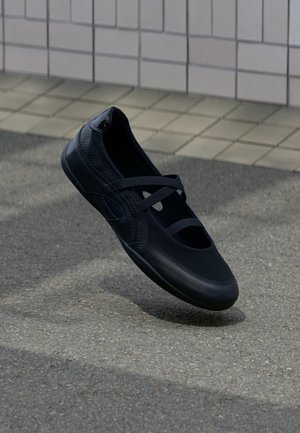 Zapato negro estilo Mary Jane con correas elásticas flotando sobre un pavimento gris texturizado, con una pared de azulejos blancos al fondo.