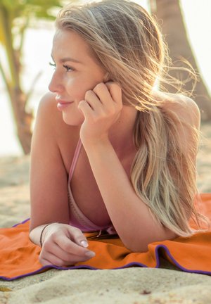 A person with long blonde hair rests on an orange towel with purple trim, touching their face, against a sandy beach background.