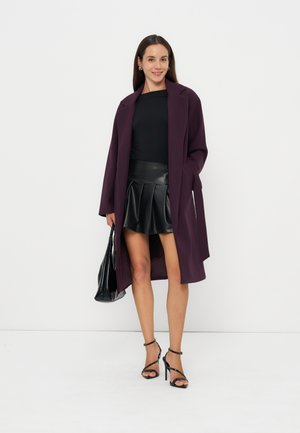 Mujer que lleva un abrigo largo morado sobre un top negro y una falda plisada de cuero negro, sosteniendo un bolso negro, de pie sobre un fondo blanco.
