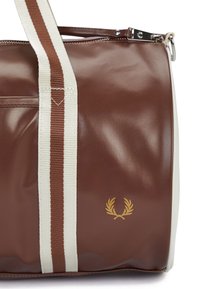 Brun vinyl duffel taske med cremefarvede stribede stofhåndtag, guld logo detaljer og lynlåslukning. Glat tekstur med blank overflade.