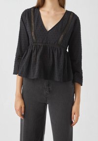 Top peplum nero con scollatura a V, texture a pois e dettagli in pizzo lungo il colletto, abbinato a pantaloni larghi grigio scuro.