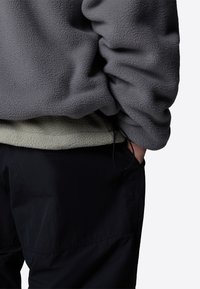 Grå fleece pullover lagdelt over lys grå fleece, med sorte bukser. Har en let tekstureret overflade, sidelomme og snøre detalje.