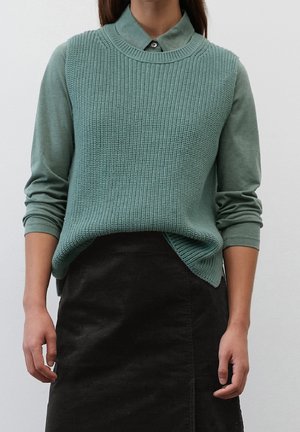 Personne portant un pull sans manches en maille verte sur une chemise verte à manches longues avec col, associé à une jupe noire taille haute.