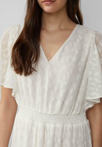 Donna che indossa un vestito bianco a scollo a V con un sottile motivo testurizzato e maniche corte a sbuffo, capelli castani che cadono sulla spalla sinistra.