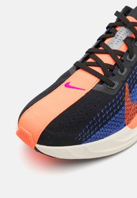 Nike løbesko med en sort mesh-overdel, der har orange og blå detaljer, teksturerede mønstre og en hvid sål. Lilla Nike swoosh-detalje.