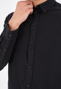 Camisa negra de botones, con tejido texturizado, mangas largas con puños abotonados, un cuello clásico y seis botones en la parte delantera.