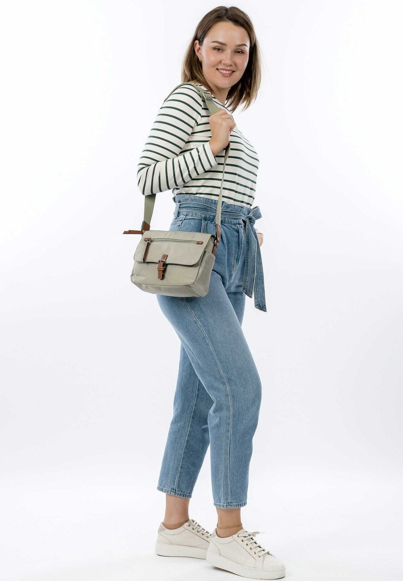 Borsa a tracolla di medie dimensioni grigia con patta, dettagli marroni e tracolla regolabile. Il modello indossa una maglietta a righe a maniche lunghe e jeans a vita alta di colore blu chiaro.