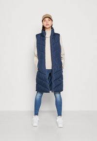 Ναυτικό μπλε επενδυμένο αμάνικο γιλέκο με ψηλό γιακά, κλείσιμο με φερμουάρ και σχέδιο σε σχήμα βέλους, σε συνδυασμό με ελαφρύ πουλόβερ και τζιν.