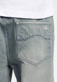 Poche arrière de jean en denim bleu clair avec des détails subtils d'usure et de rivets, partiellement recouverte par l'ourlet blanc d'une chemise.