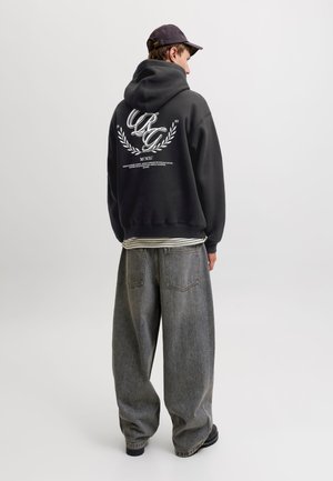 Jeune homme portant un sweat à capuche noir avec un texte graphique blanc, une casquette noire et un jean gris ample, debout devant un fond clair uni.